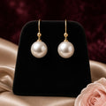 Moonlit Pearl Drop Earrings – White pearl drop earrings on gold hooks on a black velvet stand with champagne satin and blush roses (product image, invented unbranded)