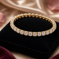 Empress Gold Tennis Bracelet – Gold tennis bracelet with white stones coiled on a black velvet cushion with champagne silk and rose petals (product image, invented unbranded)